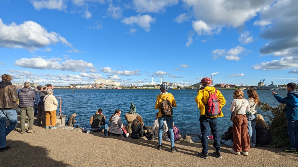 photo of about a dozen people all gathered round and taking photos of the little mermaid statue in copenhagen