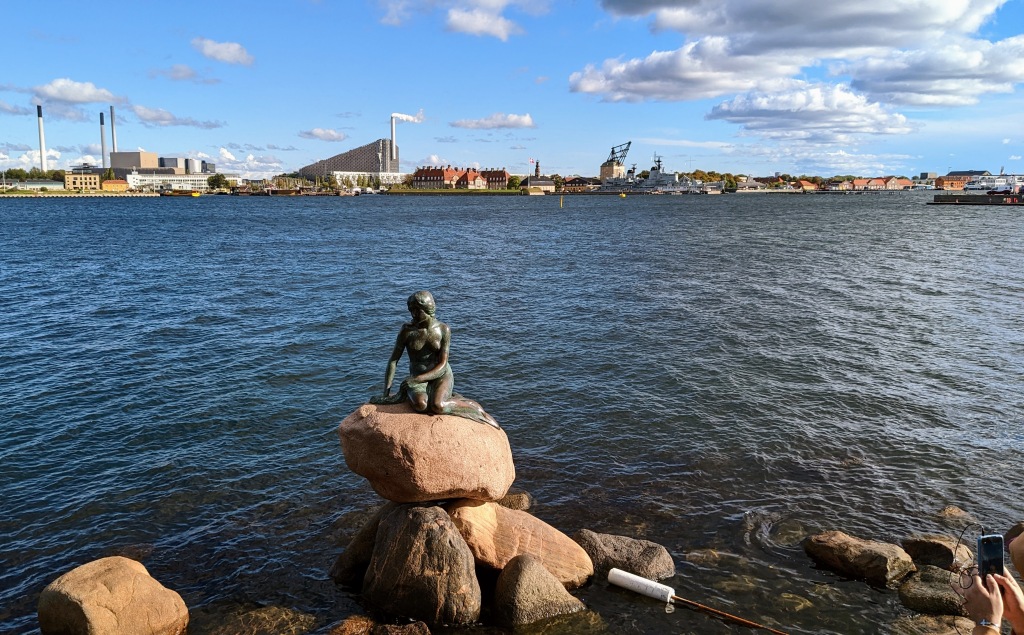 Copenhagen: the little mermaid statue perched on a rock with a blue sea background on a sunny day